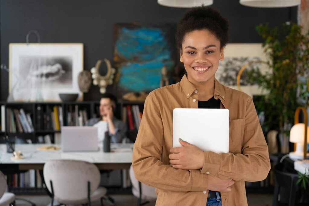 Young businesswoman with curly hair holding a laptop, smiling confidently in a modern office setting.